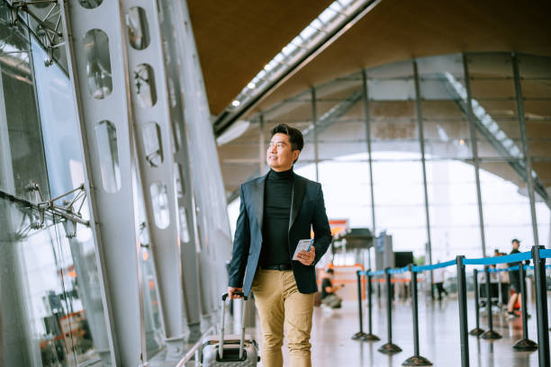 Businessman walking through airport passageway Businessman with luggage walking by window at airport terminal and watching airplanes through glass window. business journey stock pictures, royalty-free photos & images