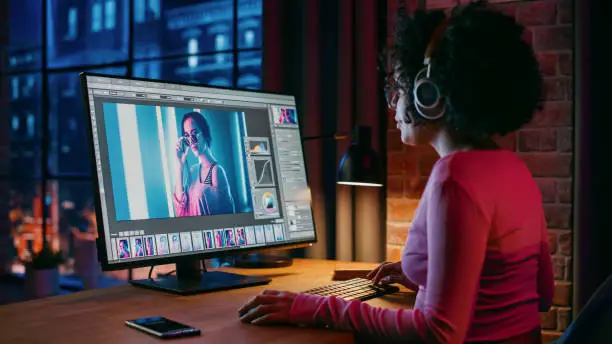 Young Creative Female in Headphones Using Computer in Stylish Loft Apartment in the Evening. Graphic Designer Working from Home, Editing Fashion Photo for Online Store. Urban View from Big Window.