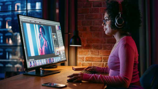 istock Young Creative Female in Headphones Using Computer in Stylish Loft Apartment in the Evening. Graphic Designer Working from Home, Editing Fashion Photo for Online Store. Urban View from Big Window. 1408847079