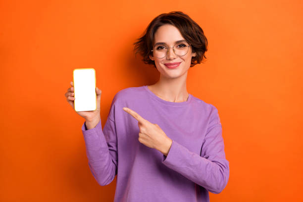 istock Photo of charming pretty girl dressed purple pullover eyewear pointing modern gadget empty space isolated orange color background 1408850218
