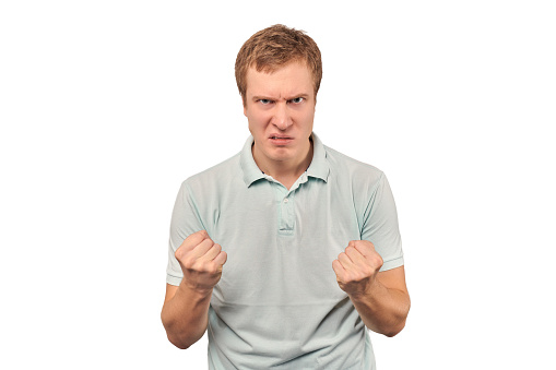 Angry young man in light grey T-shirt ready to fight with fists isolated on white background. Confident annoyed guy ready to fistfight, defense gesture, aggressive man with clenched fists
