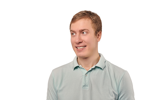 Portrait of funny young guy looking left in light grey T-shirt isolated on white background. Attractive smiling man with goodwill emotions, handsome awkward expression, male casual style