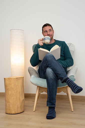 Attractive bearded man drinking a coffee while is reading a book on a modern armchair.