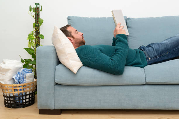 istock Side view of a attractive, bearded man lying on a sofa and reading a novel. 1408853476
