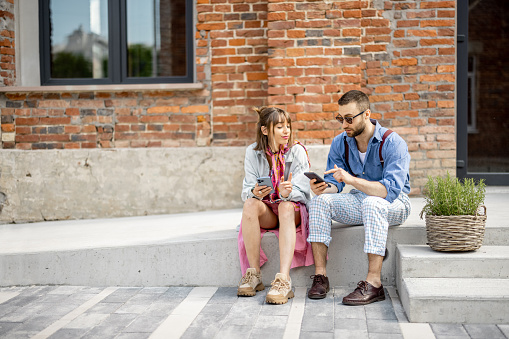 Young stylish man and woman sit together with smart phones and have some conversation near office outdoors. Modern hipsters during coffee break