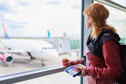 Woman standing at the airport departure area with traveling documents and coffee to go, waiting for the flight and looking through window at runway with planes