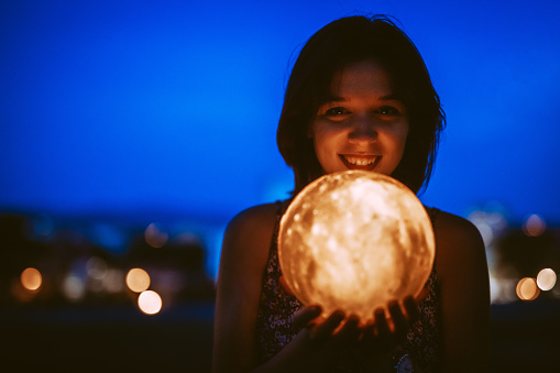 Beautiful young woman holding a lit up moon lamp at night in the city.