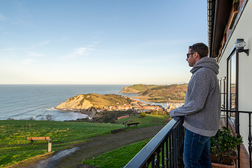Man in casual clothing standing on a balcony and looking at a sunset over zumaia, Spain.