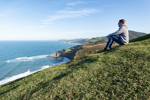Sitting man at the top of green hill looking at the flysch coastline. Basque Country destination.
