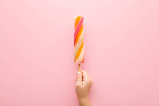 Baby girl hand holding colorful fruit ice popsicles on stick. Light pink table background. Pastel color. Closeup. Children cold sweet snack in summer. Top down view.