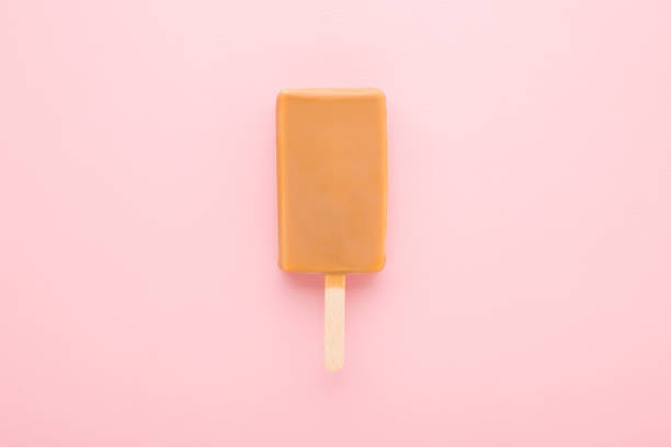 istock Light brown caramel ice cream on stick on light pink table background. Pastel color. Closeup. Top down view. 1408863187