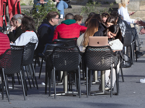 Picture of a young woman working online on a laptop, an apple macbook, surfing on the internet outdoors, from a cafe table in Bordeaux, France.