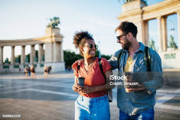 Beautiful Young Multiracial Couple Enjoying City Break Holding Coffee Cups And Wearing Backpacks Stock Photo - Download Image Now