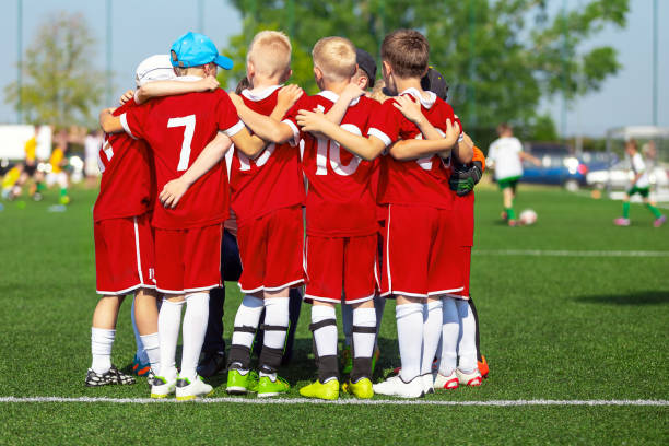 sportliche spieler drängen sich im kreis auf dem feld. motivierte jugendfußballmannschaft jubelt auf dem platz an. teamkapitän im gespräch mit einer gruppe von fußballfreunden. glückliche fußballmannschaft in roten fußballuniformen - sports uniform blue team event sports activity stock-fotos und bilder