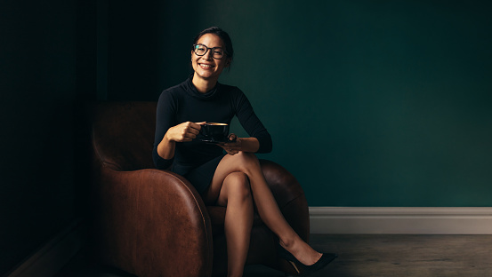Smiling woman sitting on chair and holding coffee cup. Happy asian female enjoying coffee while sitting on armchair.
