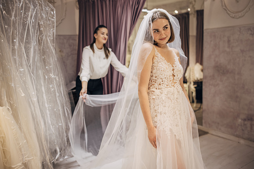 Bride wearing her wedding dress, while her female dress designer is holding a veil on dress.