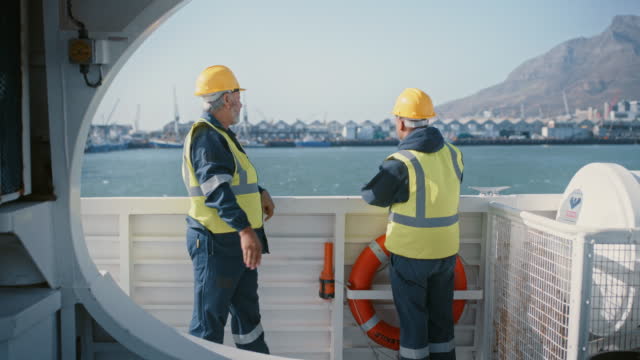 Two marine engineers from the back wearing safety uniform while discussing construction work on deck at sea. Technicians and maintenance mechanics planning to fix ship repairs and mechanical issues