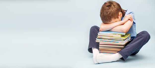 Upset schoolboy sitting with pile of school books and covers his face with hands. boy sleeping on a stack of textbooks isolated on a blue background. banner