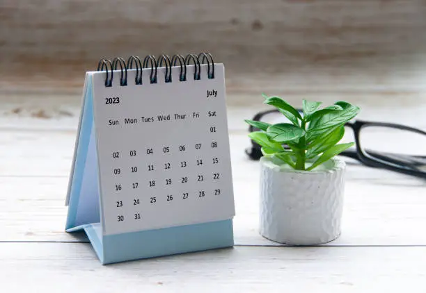 July 2023 white desk calendar on wooden table with plant and glasses background. Copy space