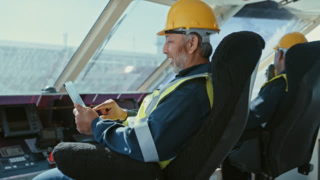 Two men on a ship on watch duty while holding a a digital tablet. Happy male mechanics with yellow vests and and safety helmets sitting in front of a merchant shipping boat overlooking the sea and using wireless technology