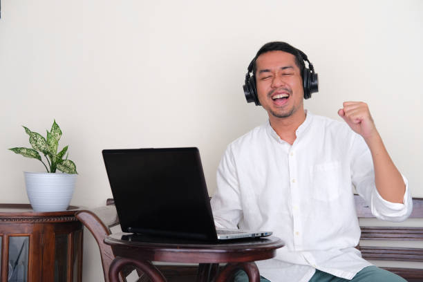 erwachsener asiatischer mann, der glück zeigt, wenn er musik hört und vor dem laptop sitzt - menari stock-fotos und bilder