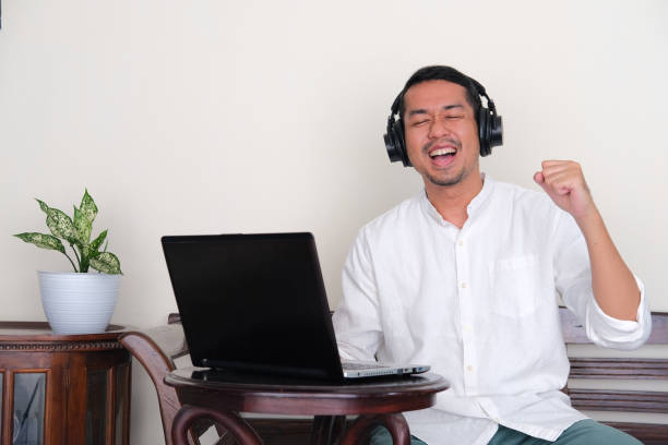 hombre asiático adulto que muestra felicidad al escuchar música y sentarse frente a la computadora portátil - menari fotografías e imágenes de stock