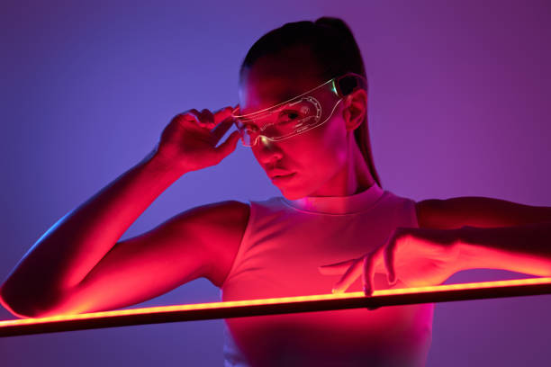 Beautiful woman in futuristic glasses leaning at the LED lamp against dark background stock photo