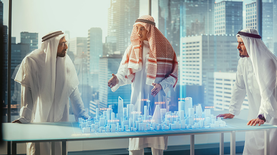 Grupo de desarrolladores de negocios inmobiliarios árabes con ropa tradicional discuten oportunidades de inversión basadas en el modelo de ciudad 3D de realidad aumentada holográfica en su oficina moderna en un rascacielos. photo
