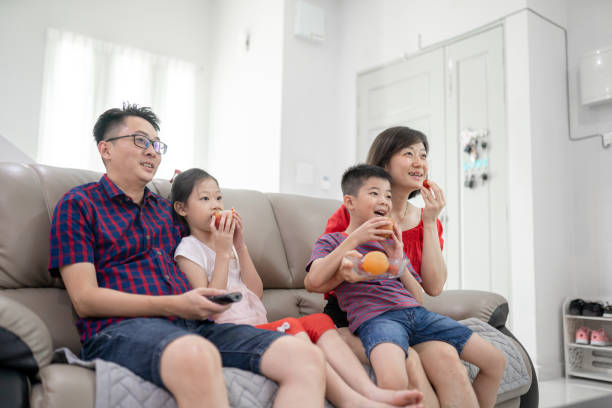 Asian Chinese family sit on sofa eating fruit in the living room laughing together watching TV Asian Chinese family sit on sofa eating fruit in the living room laughing together watching TV asian kids watching tv stock pictures, royalty-free photos & images