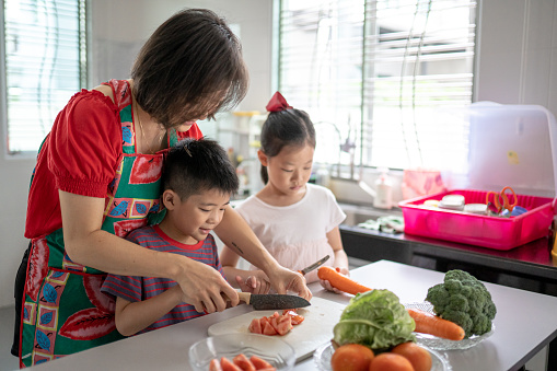 Asian Chinese mother holding kid hand teaching chopping tomato.