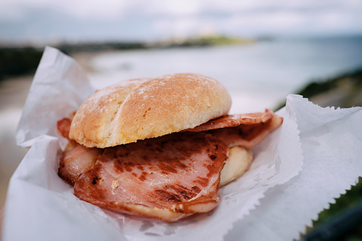 POV of a hand holding a Bacon Roll above Towan Beach, Newquay, Cornwall on a sunny June morning.