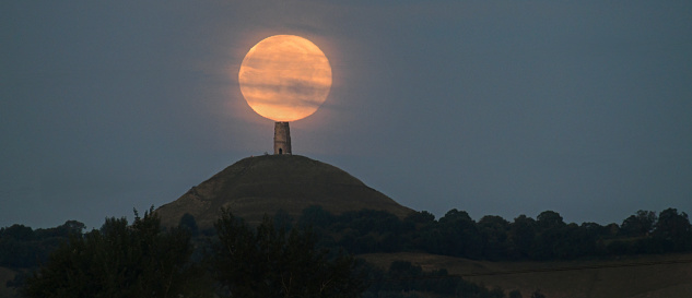 The full Supermoon appears to sit on top of Glastonbury Tor as it sets.
Glastonbury Tor is a hill near Glastonbury in the English county of Somerset. The Tor is mentioned in Celtic mythology, particularly in myths linked to King Arthur