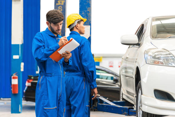 Mechanic in blue work wear uniform checks the vehicle maintenance checklist with his assistant with blur lifted car in the background. Automobile repairing service, Professional occupation teamwork. mecánico con uniforme de trabajo azul revisa la lista de verificación de mantenimiento del vehículo con su asistente con el automóvil levantado desenfoque en el fondo. servicio de reparación de automóviles, trabajo en equipo de ocupación profesional