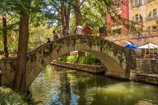 San Antonio, Texas, USA - July 3, 2018: Tourists keep cool on a bridge in the shade provided by trees on a summer day. The bridge spans the San Antonio River which runs through the city of San Antonio, Texas. It is part of a walkway that passes many restaurants and shops that line, what is called The Riverwalk, which is a popular tourist attraction.