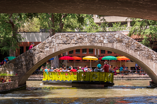 San Antonio, Texas, USA - July 1, 2018: Tourists keep cool on a summer day on a boat ride that passes under a bridge on the San Antonio River which runs through the city of San Antonio, Texas. These boat rides pass many restaurants and shops that line, what is called The Riverwalk, which is a popular tourist attraction.