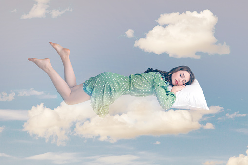 Relaxed girl in ruffle dress levitating in mid-air, sleeping on stomach lying comfortable cozy on pillow, keeping eyes closed, watching peaceful dream. collage composition on day cloudy blue sky