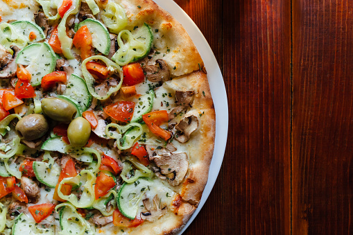 Closeup of Vegetariana Pizza with copy space on wooden table. Ingredients cheese, mushrooms, peppers, tomatoes, zucchini, olives, marinated with olive oil and garlic.