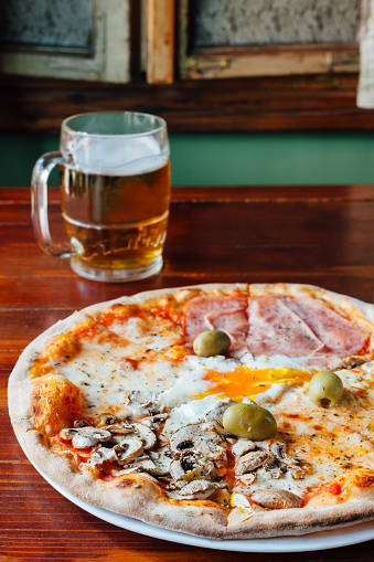 Delicious Quattro Stagioni Pizza and Beer Mug on wooden table in a rustic restaurant. Ingredients peeled tomatoes, mozzarella, cheese, ham, mushrooms, egg, olives, oregano.