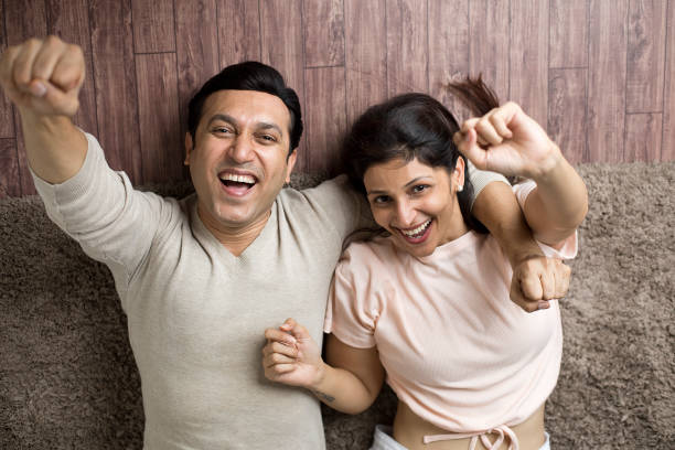 excited couple cheering while lying down with arms raised at home - couple loving lying on back carpet imagens e fotografias de stock