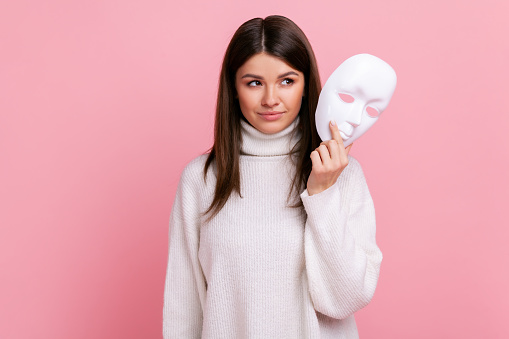Portrait of young beautiful adult female with dark hair looking away, holding in hands white mask, wearing white casual style sweater. Indoor studio shot isolated on pink background.