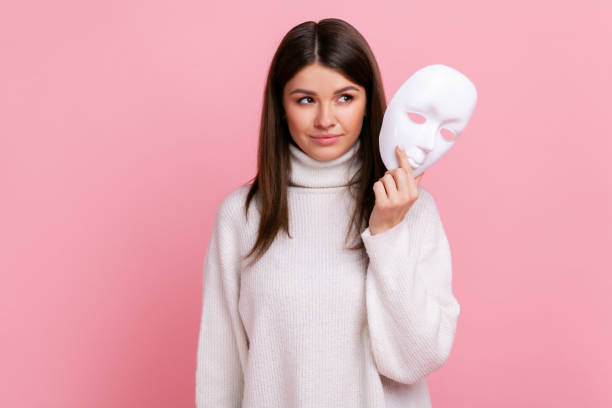 Portrait of young beautiful adult female with dark hair looking away, holding in hands white mask. Portrait of young beautiful adult female with dark hair looking away, holding in hands white mask, wearing white casual style sweater. Indoor studio shot isolated on pink background. cheesy grin stock pictures, royalty-free photos & images