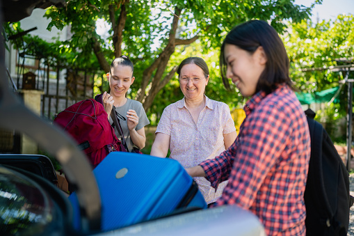 A host-mother is bringing to female tourist guests to her house for holiday home-staying by car and taking their luggages from trunk.