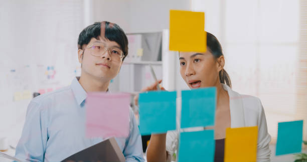 Young Asian businessman and businesswoman teamwork with formal shirt standing in front transparent whiteboard brainstorm meeting and discussing for business plan at small workplace. Young Asian businessman and businesswoman teamwork with formal shirt standing in front transparent whiteboard brainstorm meeting and discussing for business plan at small business workplace. mirror writing stock pictures, royalty-free photos & images