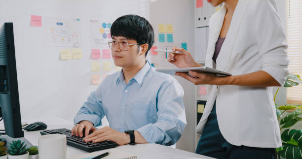 istock Close-up Young Asian businessman sit in front of computer desk look at digital tablet writing and checking for reporting work marketing and presenting for woman colleague at small workplace. 1408937507