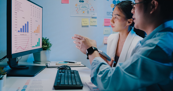 Young Asian businessman and businesswoman look at computer monitor discussing chart reporting in presentation and compare data on digital tablet at small business workplace overtime working at night.