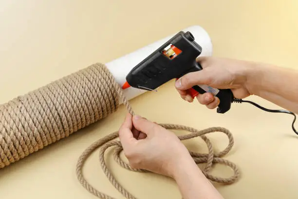 Photo of Hands hold electric hot glue gun to make rope around column for scratching post for cats