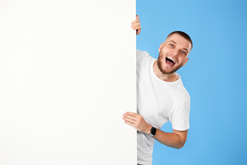friendly man peeking out from behind advertisement whiteboard with empty copy space on blue background