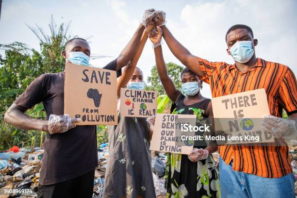 Four Young Adults Protest With Signs Against Pollution Outside An Illegal Open Landfill In Africa Stock Photo - Download Image Now