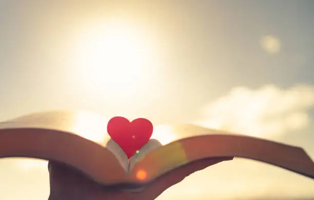 Photo of Hand holding bible book with heart against a beautiful sunset.