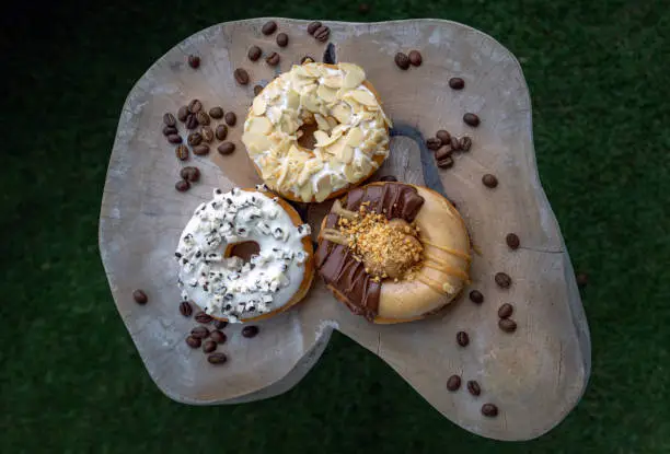 Three doughnuts covered with Cookies and creme, Almendras and Choco peanut butter on wooden cutting board in dark background. The concept of delicious glazed donuts, Top view, Space for text, Selective Focus.